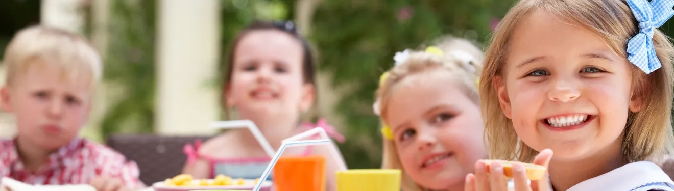 Fotogeschenke zum Geburtstag individuell gestalten Lächelnde Kinder beim Essen im Freien, mit Getränken und Snacks auf einem Tisch.
