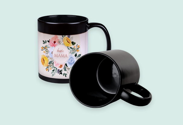 Schwarze Magic Cup, Fotomotiv erscheint erst bei Hitze Magic Cup mit floralem Motiv, das erst beim Einfüllen heißer Getränke erscheint. Daneben liegt eine komplett schwarze Tasse.