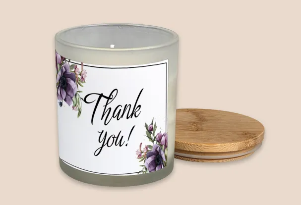 Personalisierte Duftkerze mit Bambusdeckel als Geschenkidee zur Hochzeit Duftkerze mit Deckel als Geschenkidee zur Hochzeit und Aufschrift ‚Thank you!‘, umgeben von lila Blumen auf weißem Etikett.